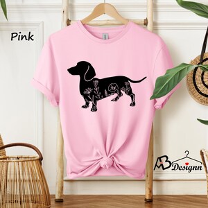 May include: Pink t-shirt featuring a black silhouette of a dachshund with floral details. The text "Pink" is visible in the top left corner of the image.