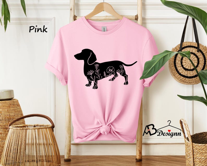 May include: Pink t-shirt featuring a black silhouette of a dachshund with floral details. The text "Pink" is visible in the top left corner of the image.