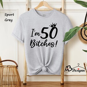 May include: A sport grey short-sleeved t-shirt featuring black text that reads I'm 50 Bitches! with a small crown graphic above the number 50. The shirt has a crew neckline and is displayed on a wooden hanger against a light-colored background.
