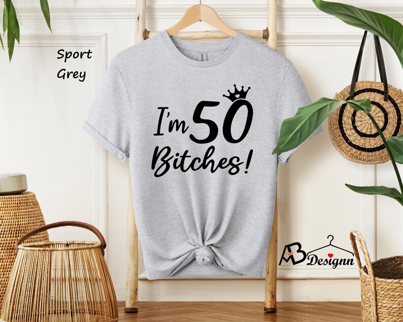 May include: A sport grey short-sleeved t-shirt featuring black text that reads I'm 50 Bitches! with a small crown graphic above the number 50. The shirt has a crew neckline and is displayed on a wooden hanger against a light-colored background.