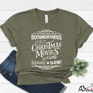 May include: Olive green t-shirt with white text that reads "IT'S EITHER SERIAL KILLER DOCUMENTARIES OR CHRISTMAS MOVIES WE EITHER SLEIGHIN' OR SLAYIN!". The shirt is folded with a pair of blue jeans.