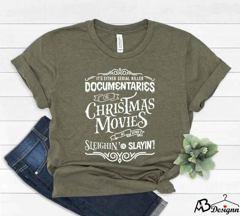 May include: Olive green t-shirt with white text that reads "IT'S EITHER SERIAL KILLER DOCUMENTARIES OR CHRISTMAS MOVIES WE EITHER SLEIGHIN' OR SLAYIN!". The shirt is folded with a pair of blue jeans.
