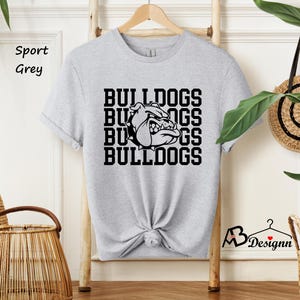Puede incluir: Una camiseta gris claro con la palabra "BULLDOGS" apilada cuatro veces en negro, con un gr&aacute;fico de bulldog en el centro. La camiseta est&aacute; colgada en una percha de madera.