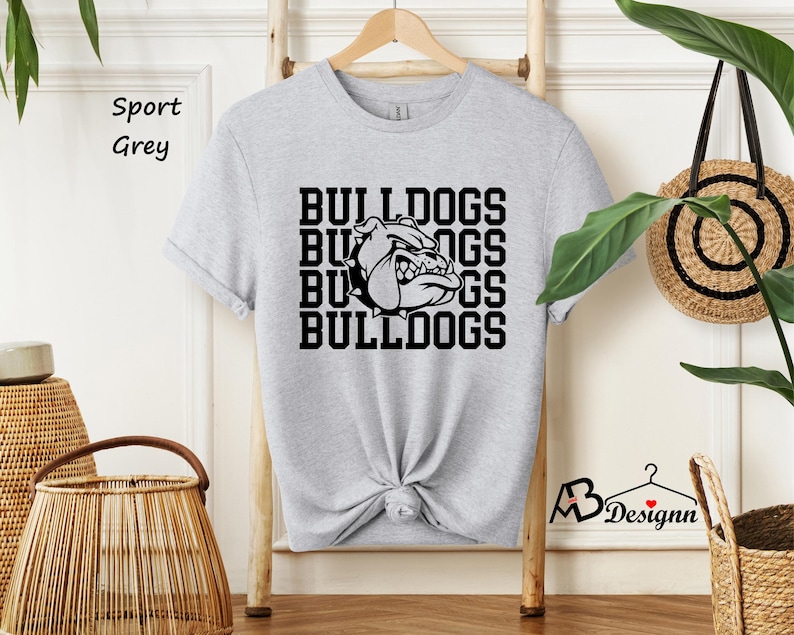 Puede incluir: Una camiseta gris claro con la palabra "BULLDOGS" apilada cuatro veces en negro, con un gr&aacute;fico de bulldog en el centro. La camiseta est&aacute; colgada en una percha de madera.