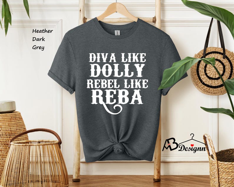Puede incluir: Una camiseta de manga corta en color gris oscuro jaspeado con texto blanco impreso en el frente que dice DIVA LIKE DOLLY REBEL LIKE REBA. La prenda tiene cuello redondo, se muestra en una percha de madera y tiene el dobladillo anudado.