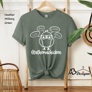 Op de afbeelding: Een groen T-shirt met een witte afbeelding van een kip met een bril en gedachtebellen met wiskundige vergelijkingen. De tekst "Mathemachicken" staat onder de kip.