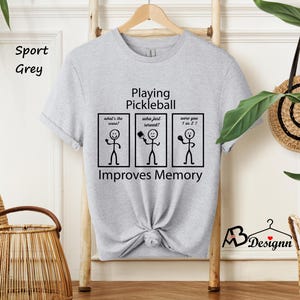 May include: A light grey t-shirt with a black graphic that says "Playing Pickleball" and has stick figures with the text "What's the score?", "Who just served?", and "Were you 1 or 2?" The graphic also says "Improves Memory".