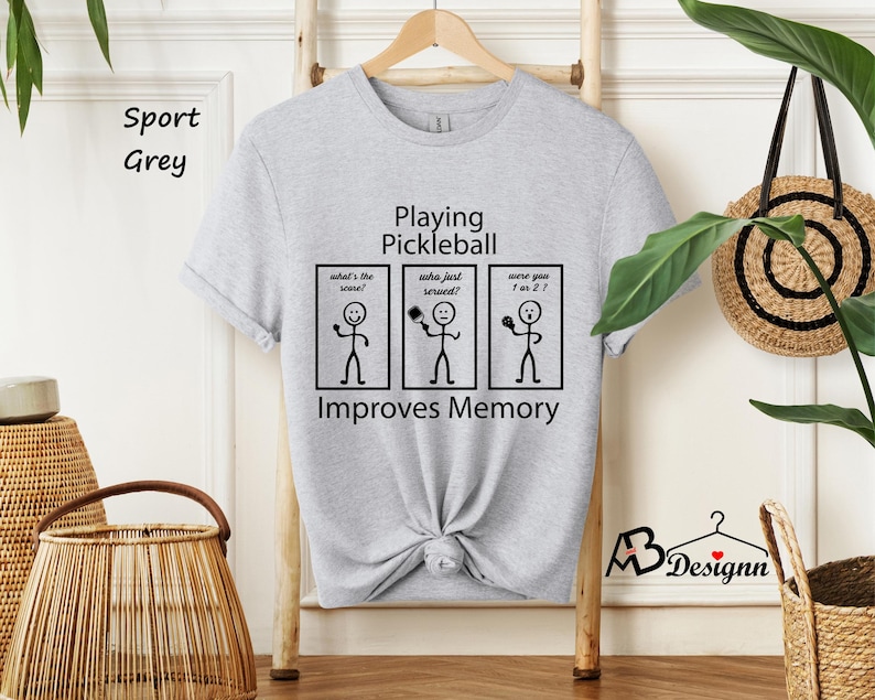 May include: A light grey t-shirt with a black graphic that says "Playing Pickleball" and has stick figures with the text "What's the score?", "Who just served?", and "Were you 1 or 2?" The graphic also says "Improves Memory".