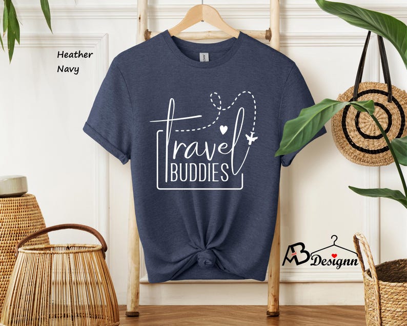 K&ouml;nnte beinhalten: Ein kurz&auml;rmeliges T-Shirt in Heather Navy mit wei&szlig;em Grafiktext, der travel BUDDIES liest. Das Design enth&auml;lt eine gestrichelte Flugbahnlinie, ein kleines Herz und die Silhouette eines Flugzeugs. Das T-Shirt h&auml;ngt an einem Holzkleiderb&uuml;gel.