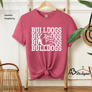 Puede incluir: Camiseta frambuesa jaspeada con la palabra "BULLDOGS" apilada cuatro veces en blanco, junto con un gr&aacute;fico de bulldog. La camiseta est&aacute; colgada en un perchero de madera. La camiseta est&aacute; anudada a la cintura.