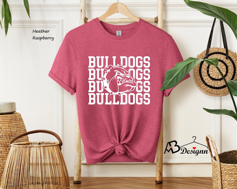 Puede incluir: Camiseta frambuesa jaspeada con la palabra "BULLDOGS" apilada cuatro veces en blanco, junto con un gr&aacute;fico de bulldog. La camiseta est&aacute; colgada en un perchero de madera. La camiseta est&aacute; anudada a la cintura.