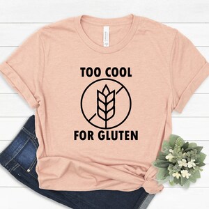 May include: A light pink t-shirt with black text that reads "Too Cool For Gluten" and a black and white graphic of a wheat stalk with a diagonal line through it.