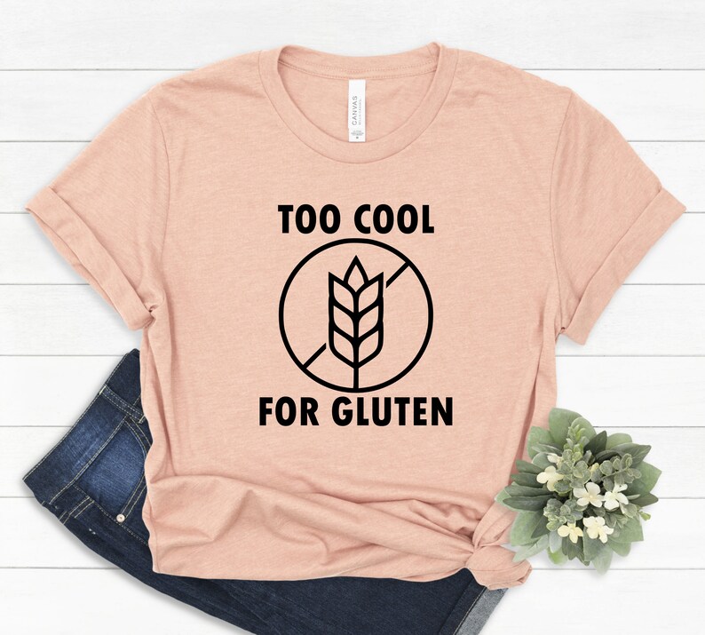 May include: A light pink t-shirt with black text that reads "Too Cool For Gluten" and a black and white graphic of a wheat stalk with a diagonal line through it.