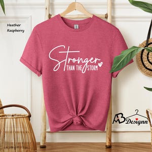 Może przedstawiać: Koszulka w kolorze malinowym heather z napisem "Stronger than the storm" w białym kolorze z małym sercem.