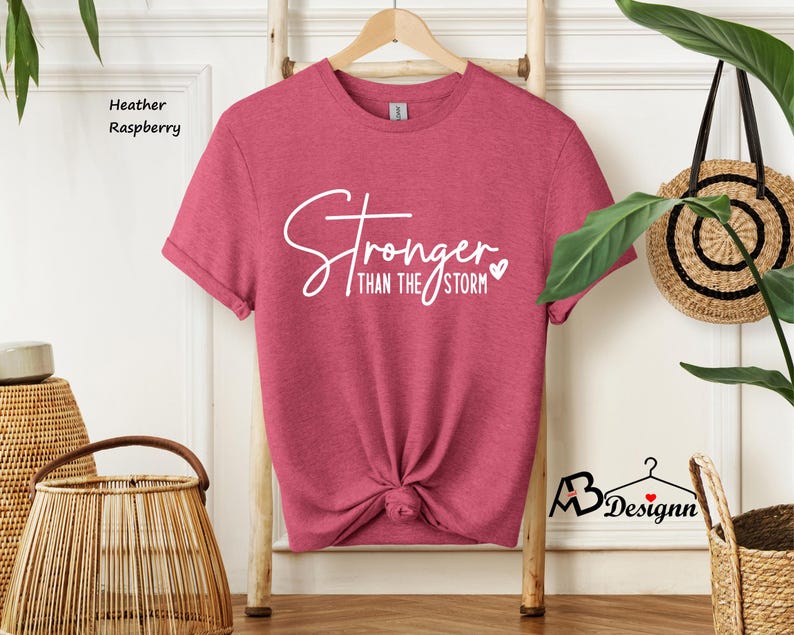 Może przedstawiać: Koszulka w kolorze malinowym heather z napisem "Stronger than the storm" w białym kolorze z małym sercem.