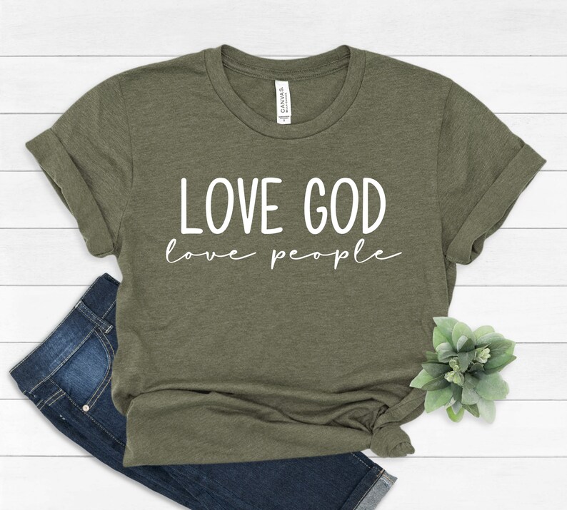 K&ouml;nnte beinhalten: Olivgr&uuml;nes T-Shirt mit dem wei&szlig;en Text "LOVE GOD love people". Das Shirt ist mit einer Jeans und einer kleinen gr&uuml;nen Pflanze gefaltet. Das T-Shirt besteht aus einem weichen Material.