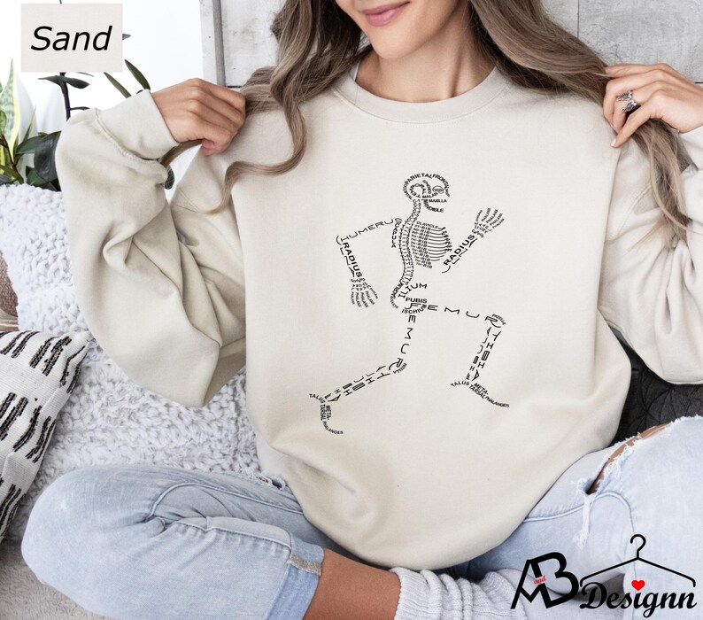 Peut inclure: Sweat-shirt couleur sable avec un motif noir d'un squelette courant avec des os &eacute;tiquet&eacute;s. Le texte "Sand" est dans le coin sup&eacute;rieur gauche. Le sweat-shirt est de couleur neutre et a une coupe d&eacute;contract&eacute;e.