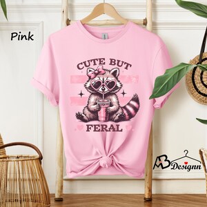 Op de afbeelding: Roze T-shirt met een afbeelding van een wasbeer die een zonnebril draagt en een roze drankje vasthoudt. De tekst "Cute But Feral" staat op het T-shirt gedrukt.