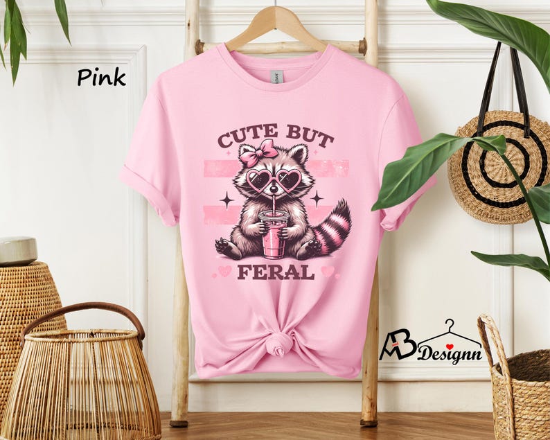 Op de afbeelding: Roze T-shirt met een afbeelding van een wasbeer die een zonnebril draagt en een roze drankje vasthoudt. De tekst "Cute But Feral" staat op het T-shirt gedrukt.