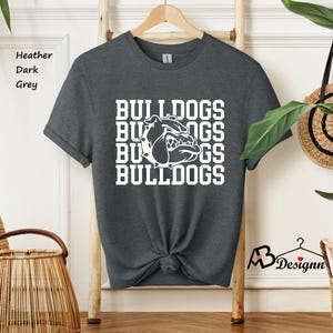 Puede incluir: Camiseta gris oscuro jaspeado con la palabra "BULLDOGS" apilada verticalmente en blanco, con un gr&aacute;fico de bulldog en el centro. La camiseta est&aacute; colgada de una percha de madera.