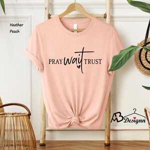 May include: Heather peach-colored t-shirt with the words "PRAY WAIT TRUST" in black script. The shirt is hanging on a wooden hanger against a white wall. A woven basket and a decorative wall hanging are also visible.