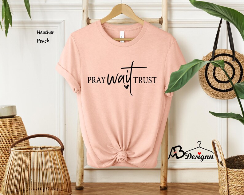 May include: Heather peach-colored t-shirt with the words "PRAY WAIT TRUST" in black script. The shirt is hanging on a wooden hanger against a white wall. A woven basket and a decorative wall hanging are also visible.
