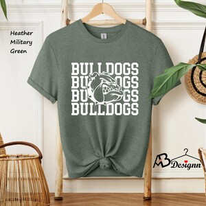Puede incluir: Camiseta verde militar jaspeada con la palabra "BULLDOGS" apilada verticalmente en blanco, junto con un gr&aacute;fico de bulldog blanco. La camiseta est&aacute; colgada de una percha de madera.