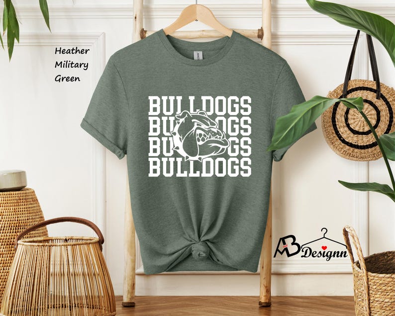 Puede incluir: Camiseta verde militar jaspeada con la palabra "BULLDOGS" apilada verticalmente en blanco, junto con un gr&aacute;fico de bulldog blanco. La camiseta est&aacute; colgada de una percha de madera.