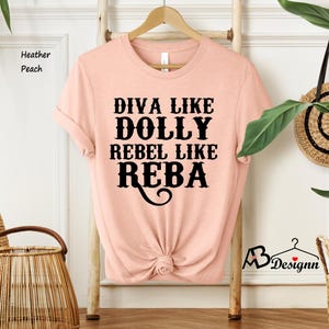Puede incluir: Una camiseta de manga corta color melocot&oacute;n jaspeado con cuello redondo. La parte delantera presenta un texto en negro que dice DIVA LIKE DOLLY REBEL LIKE REBA en una fuente estilizada. La parte inferior est&aacute; recogida en un peque&ntilde;o nudo.