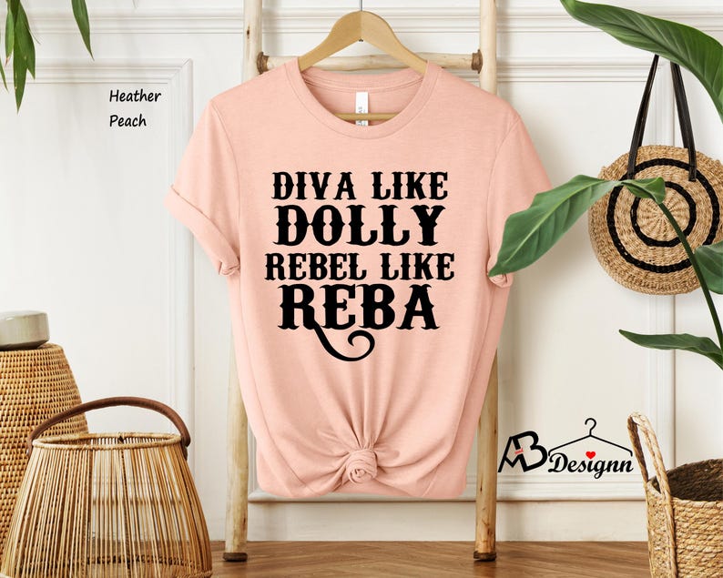 Puede incluir: Una camiseta de manga corta color melocot&oacute;n jaspeado con cuello redondo. La parte delantera presenta un texto en negro que dice DIVA LIKE DOLLY REBEL LIKE REBA en una fuente estilizada. La parte inferior est&aacute; recogida en un peque&ntilde;o nudo.
