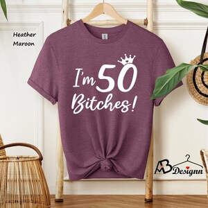 May include: A heather maroon short-sleeve t-shirt featuring white screen-printed text that reads I'm 50 Bitches! with a small crown icon above the number 50. The shirt is displayed on a wooden hanger and has a knotted hem at the bottom center.