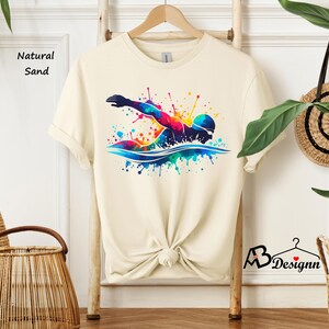 May include: A cream colored t-shirt with a colorful graphic of a swimmer in a watercolor style. The text "Natural Sand" is visible in the top left corner of the image.