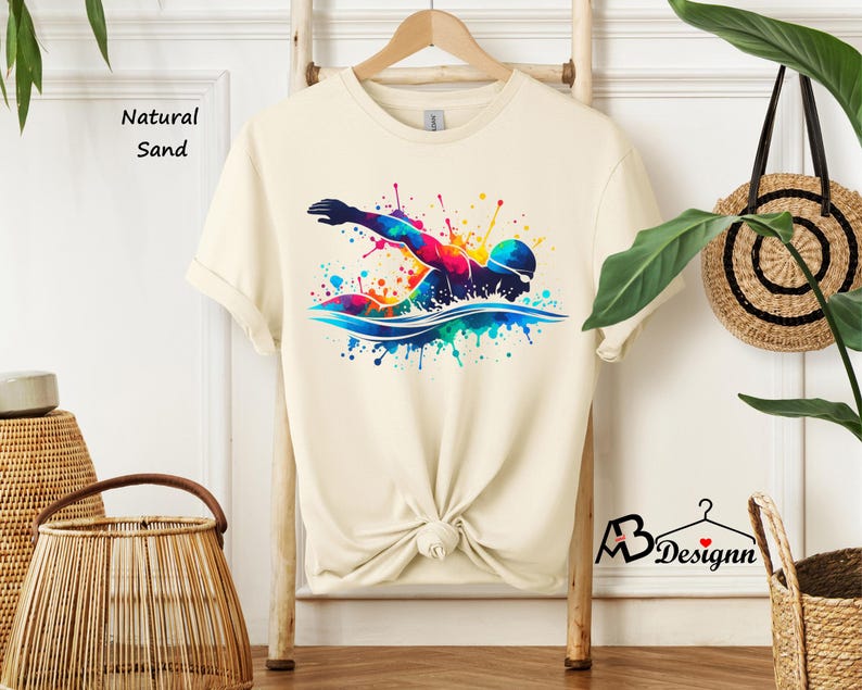 May include: A cream colored t-shirt with a colorful graphic of a swimmer in a watercolor style. The text "Natural Sand" is visible in the top left corner of the image.