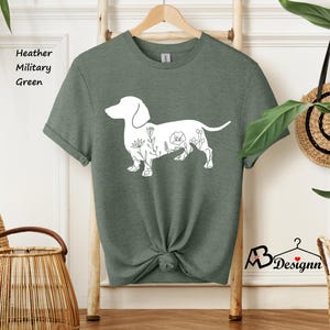 May include: A heather military green t-shirt featuring a white outline of a dachshund with floral details. The text "Heather Military Green" is visible in the top left corner of the image.