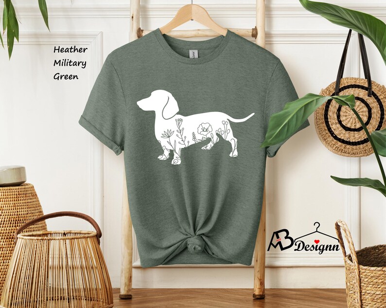 May include: A heather military green t-shirt featuring a white outline of a dachshund with floral details. The text "Heather Military Green" is visible in the top left corner of the image.