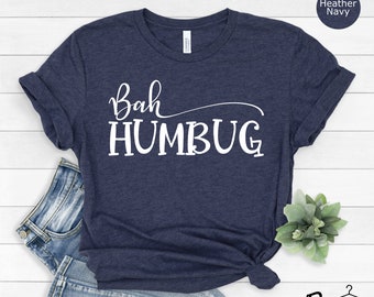 May include: Heather navy blue t-shirt with the phrase "Bah Humbug" in white script. The shirt is folded at the bottom and paired with blue jeans. The words "Heather Navy" are in a circle in the upper right corner.