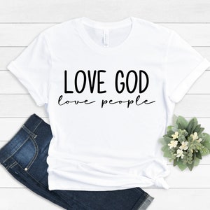 K&ouml;nnte beinhalten: Wei&szlig;es T-Shirt mit dem schwarzen Text "LOVE GOD love people". Das Shirt wird mit blauen Jeans und einem kleinen Blumenarrangement kombiniert. Das T-Shirt besteht aus einem weichen Material.