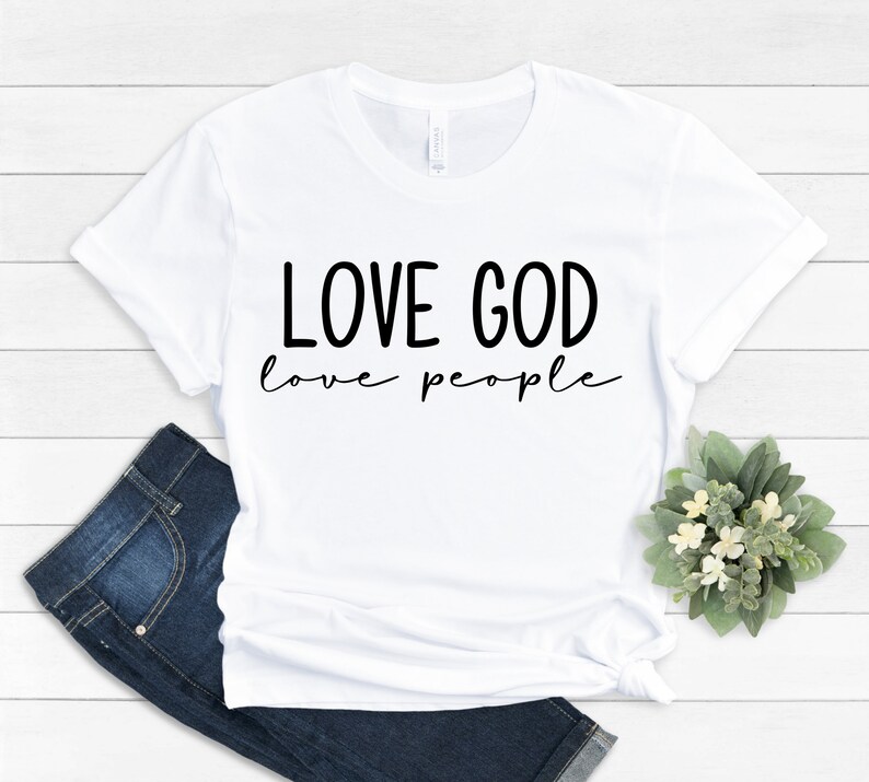 K&ouml;nnte beinhalten: Wei&szlig;es T-Shirt mit dem schwarzen Text "LOVE GOD love people". Das Shirt wird mit blauen Jeans und einem kleinen Blumenarrangement kombiniert. Das T-Shirt besteht aus einem weichen Material.