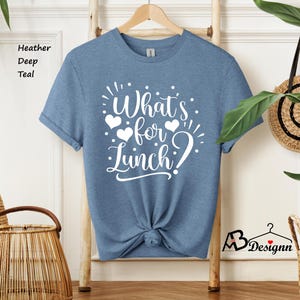 K&ouml;nnte beinhalten: Ein T-Shirt in der Farbe Heide-Tiefe Teal mit dem Text "What's for Lunch?" in Wei&szlig;. Der Text ist von drei wei&szlig;en Herzen umgeben.