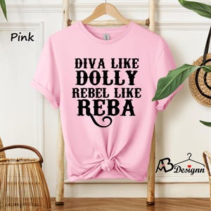 Puede incluir: Una camiseta de manga corta de color rosa claro con el texto en negro que dice DIVA LIKE DOLLY REBEL LIKE REBA. La prenda tiene cuello redondo y est&aacute; anudada en la parte inferior central. La tela es de color rosa claro liso con letras negras en negrita.