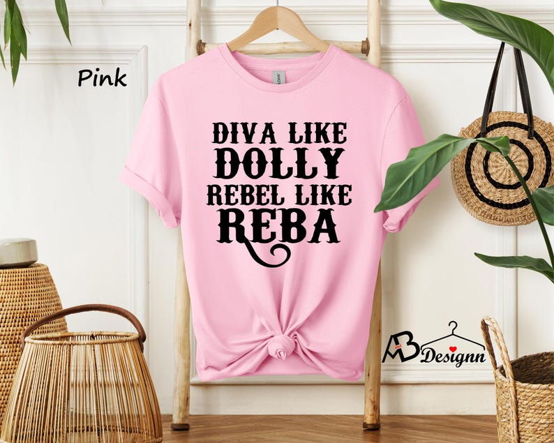 Puede incluir: Una camiseta de manga corta de color rosa claro con el texto en negro que dice DIVA LIKE DOLLY REBEL LIKE REBA. La prenda tiene cuello redondo y est&aacute; anudada en la parte inferior central. La tela es de color rosa claro liso con letras negras en negrita.