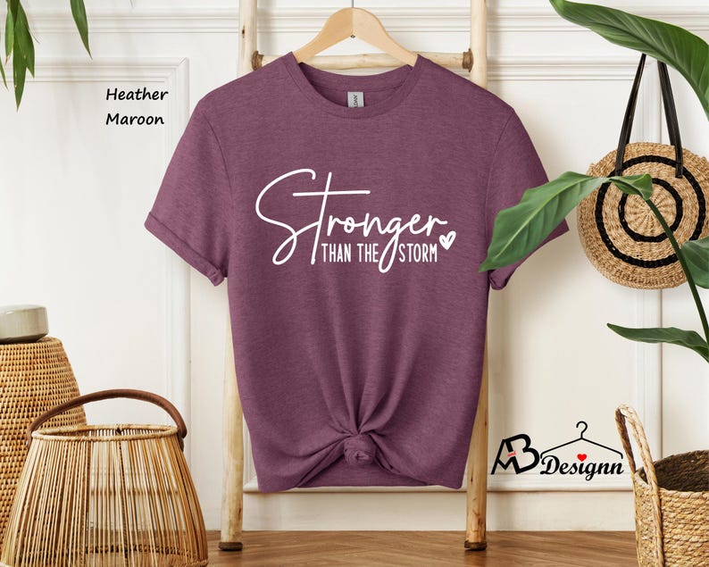 Może przedstawiać: Koszulka w kolorze brązowym heather z napisem "Stronger than the storm" w białym kolorze z małym sercem.