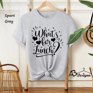 K&ouml;nnte beinhalten: Ein graues T-Shirt mit dem Text "What's for Lunch?" in schwarzer Schrift. Der Text ist von kleinen schwarzen Herzen umgeben.