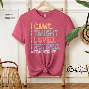 Może przedstawiać: Koszulka w kolorze wrzosu malinowego z napisem "I CAME. I TAUGHT. I LOVED. I RETIRED. #TEACHERLIFE" w kolorowych literach. Koszulka jest zawiązana w pasie i wisi na drewnianym wieszaku.