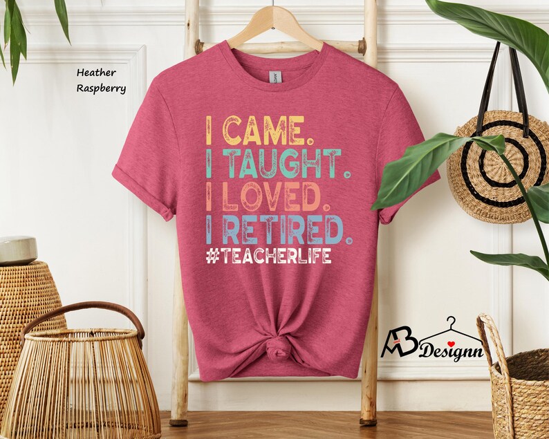Może przedstawiać: Koszulka w kolorze wrzosu malinowego z napisem "I CAME. I TAUGHT. I LOVED. I RETIRED. #TEACHERLIFE" w kolorowych literach. Koszulka jest zawiązana w pasie i wisi na drewnianym wieszaku.