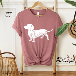 May include: A heather mauve t-shirt featuring a white outline of a dachshund with floral details. The shirt is folded and displayed on a wooden hanger.