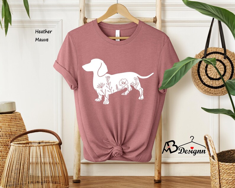 May include: A heather mauve t-shirt featuring a white outline of a dachshund with floral details. The shirt is folded and displayed on a wooden hanger.