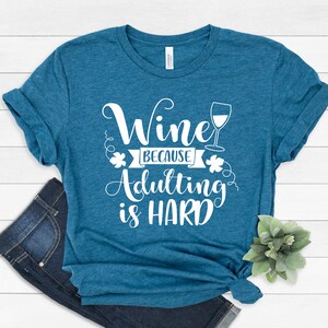 Pu&ograve; includere: T-shirt color teal con la scritta bianca "Wine because adulting is HARD". Un'immagine di un bicchiere da vino &egrave; accanto alla parola "Wine". La maglietta &egrave; piegata e annodata in basso, con un paio di jeans blu visibili.