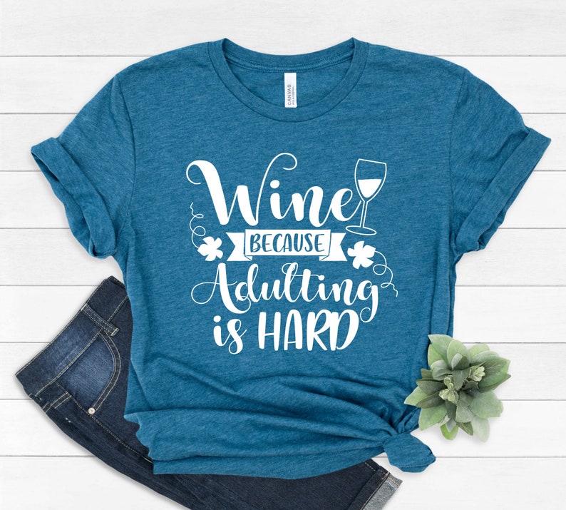 Pu&ograve; includere: T-shirt color teal con la scritta bianca "Wine because adulting is HARD". Un'immagine di un bicchiere da vino &egrave; accanto alla parola "Wine". La maglietta &egrave; piegata e annodata in basso, con un paio di jeans blu visibili.