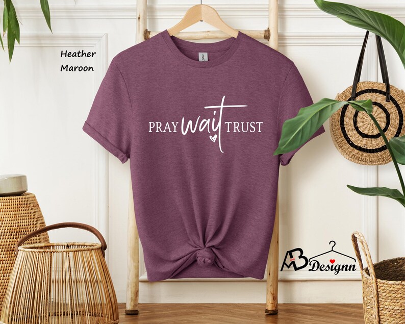May include: Heather maroon t-shirt with the words "PRAY wait TRUST" in white script. The shirt is hanging on a wooden hanger. The shirt is knotted at the waist.